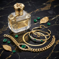 Perfume bottle labeled 'Serpentine's Heart' with gold jewelry and emeralds on a dark marble surface