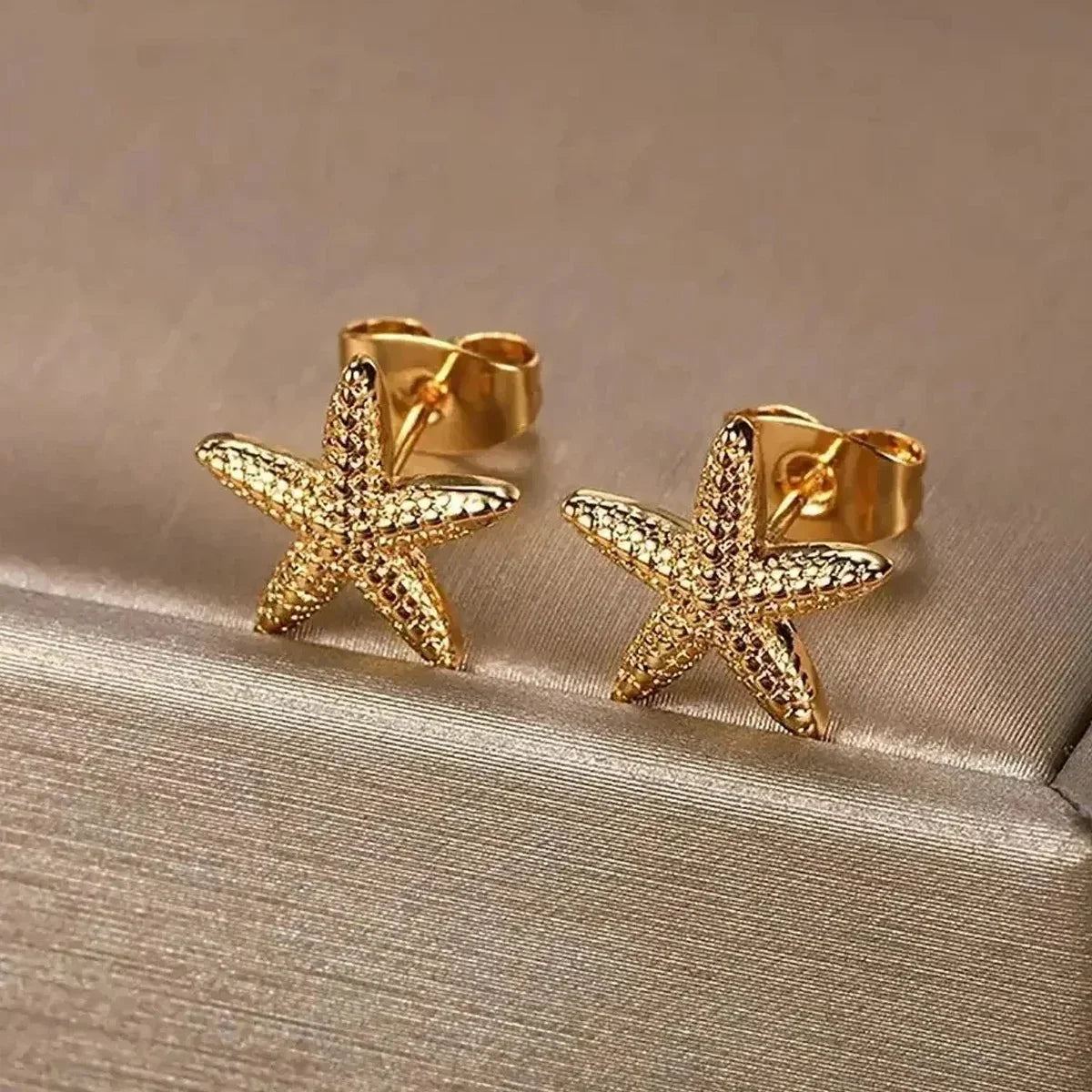 Gold starfish stud earrings with textured details on a beige fabric surface